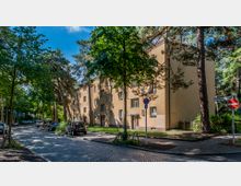 Wohnstraße mit mehrstöckigem Wohngebäude in gelblicher Farbe, umgeben von hohen Bäumen. Die gepflasterte Straße ist von parkenden Autos gesäumt, und ein Straßenschild markiert den Hartmannsweilerweg.