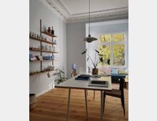 Helles Wohnzimmer mit hohen Decken und Stuckverzierungen, ausgestattet mit einem Holzboden, einem weißen Tisch, einer Regalwand mit dekorativen Gegenständen und einem großen Fenster mit Blick auf gelb belaubte Bäume. Im Hintergrund stehen ein buntes Sofa mit Kissen und eine Zimmerpflanze.