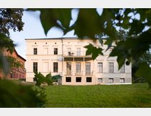 Helles, zweistöckiges klassizistisches Gebäude mit symmetrischer Fassade, großen Fenstern und einem Balkon auf der Rückseite, umgeben von einer grünen Wiese und eingebettet in Bäume. Links im Bild ist ein älteres rotes Ziegelgebäude zu sehen.