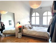 Schlafzimmer mit einem großen Bett, flankiert von einem Nachttisch mit Blumenvase und einer Wandlampe. Der Raum hat helle Wände, einen Holzboden, drei hohe Bogenfenster und einen großen Spiegel an der linken Wand, der den Raum reflektiert. Ein Kleiderständer mit Kleidung steht rechts und links des Raumes.