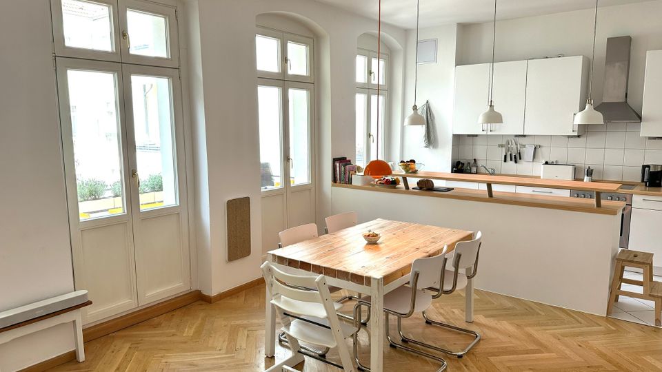 Heller Wohnküchenbereich mit hohen, bogenförmigen Fenstern, Fischgrät-Parkettboden und einem Holztisch mit mehreren Stühlen im Vordergrund. Im Hintergrund eine moderne weiße Küchenzeile mit Fliesenrückwand, Dunstabzugshaube und pendelnden Deckenlampen über einer langen Arbeitsfläche.