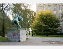 Eine bronzene Reiterstatue auf einem steinernen Sockel steht in einem Park mit Bäumen und herbstlichem Laub, vor einem hellen, mehrstöckigen Gebäude mit großen Fenstern. Auf dem Sockel ist rotes Graffiti angebracht.