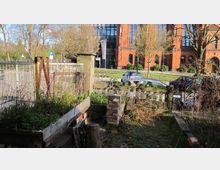 Ein kleiner Garten mit Hochbeeten und einer Holzlattenumzäunung im Vordergrund. Im Hintergrund stehen parkende Autos, eine Straße und ein großes, historisches Backsteingebäude mit hohen Fenstern.