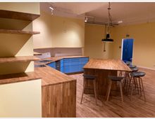 Offene Küche mit gelben Wänden, Holzarbeitsflächen und blauen Unterschränken. Im Zentrum steht ein großer Holztisch mit mehreren Barhockern, darüber hängt eine schwarze Pendelleuchte.