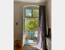 Ein heller Wohnraum mit einem offenen, hohen Fenster mit Rundbogen, das auf einen kleinen Balkon führt. Der Balkon ist mit einem blauen runden Tisch, einem gemütlichen Stuhl mit Fell und Pflanzen dekoriert, umgeben von dichtem, grünem Baumwuchs.