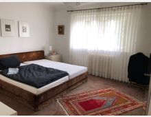 Ein Schlafzimmer mit einem Doppelbett mit braunem Stoffrahmen und dunkler Bettwäsche, einem Nachttisch mit Lampe und Vorhängen, die vor einem großen Fenster hängen. Auf dem Teppichboden liegt ein farbenfroher orientalischer Teppich, und an der Wand hängen einige Bilder.