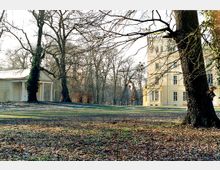 Parklandschaft mit kahlen Bäumen im Herbst oder Winter, einem kleinen klassizistischen Pavillon links und einem großen, gelb gestrichenen historischen Gebäude mit symmetrischen Fenstern im Hintergrund.