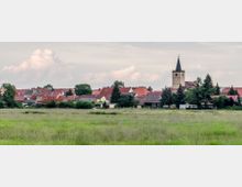 Kleinstadtlandschaft mit einer Kirche im Hintergrund, deren Turm mit einem spitzen Dach und einer Uhr versehen ist, umgeben von roten und braunen Dächern sowie Bäumen; im Vordergrund liegt eine weite grüne Wiese.