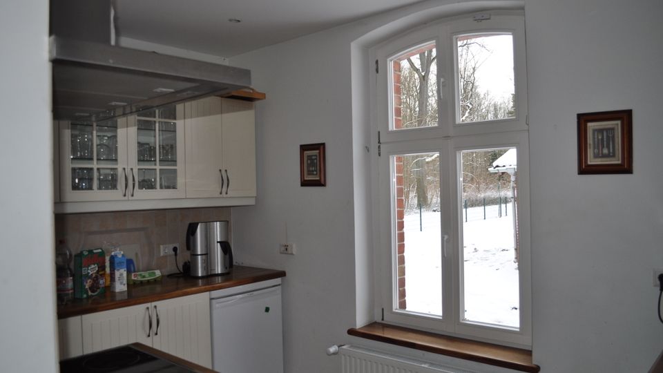 Helle Küche mit weißen Einbauschränken, Holzarbeitsplatten und einem großen, weiß gerahmten Fenster, das den Blick auf eine verschneite Außenlandschaft freigibt. Eine Dunstabzugshaube, kleinere Dekorationsbilder und ein Heizkörper unterhalb des Fensters ergänzen die Einrichtung.