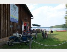 Holzgebäude mit einem Banner für Boots- und Fahrradverleih, davor geparkte Fahrräder und ein Motorrad. Im Hintergrund eine Wiese mit Sitzgelegenheiten, ein See mit Booten und Bäumen am Ufer.