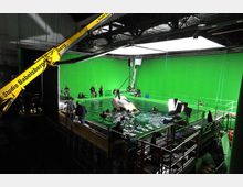 Filmstudio mit einer Wasserbühne, beleuchtet von Studiolampen. Der Hintergrund zeigt eine große grüne Leinwand (Greenscreen), vor der Personen und ein ins Wasser eintauchendes Auto für eine Filmaufnahme arrangiert sind.