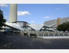 Großer öffentlicher Platz mit Springbrunnen im Vordergrund, modernen Sitzbänken und einer weißen, futuristisch gestalteten Gebäudestruktur im Hintergrund, die als Hauptelement dient. Oberhalb ragt ein Ausschnitt eines großen, runden Fernsehturms empor, umgeben von wolkenlosem Himmel.