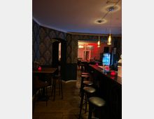 Barraum mit gedämpfter Beleuchtung und stilvoll gemusterten Tapeten. Im Vordergrund befinden sich Hocker und eine lange Theke mit roten Kerzen, im Hintergrund sind weitere Sitzgelegenheiten und ein beleuchteter Getränkekühlschrank sichtbar.