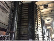 Industrieanlage mit großen vertikalen Metalltanks, die durch Gürtel und Nieten verstärkt sind, in einem gealterten Gebäude mit abgenutzten Wänden und einer grob verarbeiteten Decke. Im Vordergrund ist ein Metallzaun sichtbar.