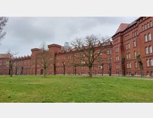 Großes historisches Backsteingebäude mit zahlreichen weißen Bogenfenstern und Ziertürmen, umgeben von einer Wiese mit vereinzelten, kahlen Bäumen unter einem grauen Himmel.