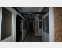 Ein dunkler, verlassen wirkender Flur mit abgenutzten Wänden und eingebauten Holz- und Glaspaneelen. Deckenrohre und unverkleidete Betondecken sind sichtbar, der Raum wirkt unbeleuchtet und heruntergekommen.