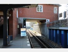 Bahnhof mit überdachtem Bahnsteig aus Beton und Backsteinwänden, einem Gleis mit Kurve, einem grünen Signalsystem und der Markierung „4a“. Im Hintergrund ist eine Überführung sowie eine Stützmauer mit blauen Elementen zu sehen.