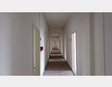 Langer, schmaler Flur in einem Gebäude mit weißen Wänden und geöffneter Tür auf beiden Seiten. An der Wand hängt ein roter Feuermelder, und der Boden ist mit einem Teppich bedeckt.