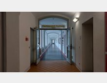 Ein heller Flur in einem Gebäude, beschriftet mit „Fachbereich Technik“ über einer offenen Doppeltür. Der Gang hat weiße Wände, ein grauer Boden und rote Streifen entlang der Wände; Fenster und Heizkörper befinden sich auf der linken Seite, Schaukästen rechts.