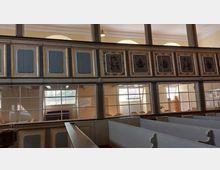 Innenraum einer historischen Kirche mit bemalten Holzvertäfelungen, verziert mit goldenen Rahmen und Wappenmotiven. Im unteren Bereich sind Fenster, die einen Blick in einen angrenzenden Gemeinderaum mit Stühlen und großen Fenstern erlauben.