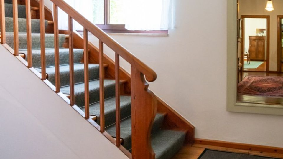 Heller Innenflur mit Holztreppe und Teppichstufen, weiß gestrichenen Wänden und Holzdielenboden. Ein Fenster mit halbtransparenten Vorhängen sowie ein großer Wandspiegel, der einen angrenzenden Raum zeigt, sind zu sehen.