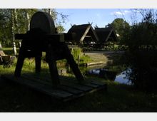Eine ländliche Szene mit einem alten Schleifstein im Vordergrund, umgeben von Gras und Bäumen. Im Hintergrund sind mehrere Holzhütten mit Reetdächern und eine kleine Terrasse am Ufer eines Teiches zu sehen.