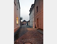 Eine schmale gepflasterte Gasse mit einfachen, zweistöckigen Wohnhäusern auf beiden Seiten, einige Fenster erhellt. Am Ende der Gasse sind weitere Gebäude mit Ziegeldächern sichtbar, umgeben von einer ruhigen Abenddämmerung.
