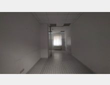 Leerer Raum mit weißen Fliesenboden und weißen Wänden, ein Fenster mit vertikalen Jalousien befindet sich am Ende des Raumes. Die abgehängte Decke hat integrierte Lichtpaneele, und an der rechten Wand sind Fliesenelemente sichtbar.