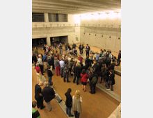 Eine Gruppe von Menschen steht in einem offenen Raum mit Betonwänden und einem kargen, minimalistischen Design. Der Raum hat klare architektonische Linien, sichtbare Metallbefestigungen und eine Decke mit mehreren Lichtquellen.