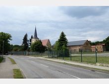 Straßenansicht mit einer gepflasterten Straße, einem Gehweg und einem eingezäunten Gelände. Im Hintergrund befinden sich eine Kirche mit spitzem Turm und roten Ziegeldächern sowie mehrere Nebengebäude aus Backstein und Holz.