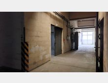 Ein Industriekorridor mit Betonwänden und einem roh gestalteten Boden. Zu sehen sind Metalltüren, freiliegende Kabel an den Wänden und ein heller Bereich mit einem großen, halbtransparenten Fenster am Ende.