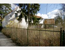 Einfache weiße Gebäude mit rotem Ziegeldach hinter einem Metallzaun, umgeben von Bäumen und Sträuchern. Ein Schild an der rechten Seite trägt die Aufschrift "Bootshaus Weller" mit zusätzlichen Kontaktdetails.