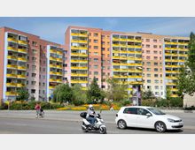 Mehrstöckiges Wohngebäude mit farbigen Fassaden in Rosa, Orange und Beige sowie zahlreichen Balkonen mit gelben Brüstungen. Im Vordergrund eine Straße mit vorbeifahrenden Fahrzeugen und eine bepflanzte Grünfläche mit Bäumen und Blumen.