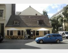 Kleines gelbes Gebäude mit einem roten Ziegeldach und zwei Dachgaubenfenstern, das ein Café oder Restaurant beherbergt. Vor dem Gebäude stehen Tische mit Stühlen unter Sonnenschirmen, und ein blauer Kombi parkt auf der Straße davor.