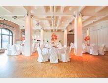 Großer Veranstaltungsraum mit heller, moderner Einrichtung, Holzboden und weiß gestrichenen Säulen. Runde Tische mit weißen Tischdecken und Stuhlhussen sind festlich gedeckt, dekoriert mit roten Kunstblumen; eine Ziegelwand und große Fenster mit Rundbögen sind im Hintergrund zu sehen.