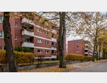 Mehrstöckige Wohngebäude aus rotem Backstein mit weißen Balkonen, umgeben von Bäumen und Hecken. Der Gehweg ist mit Herbstlaub bedeckt, und die Szene zeigt eine ruhige Wohnstraße.