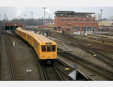 Gelbes U-Bahn-Zugmodell fährt auf einem Gleis in einem Bahnbetriebswerk, umgeben von mehreren Gleisen, Weichen und Signalen; im Hintergrund stehen ein modernes Backsteingebäude und weitere Züge.