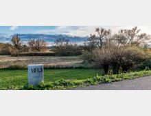 Eine ländliche Landschaft mit einer grünen Wiese, vereinzelt stehenden Bäumen und Büschen sowie einem Kilometerstein mit der Aufschrift "101,3" im Vordergrund. Im Hintergrund erstreckt sich ein bewaldetes Gebiet unter einem leicht bewölkten Himmel.