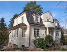 Zweistöckiges Wohnhaus im Landhausstil mit hellbeiger Fassade, dunkelbraunem Dach und mehreren Gauben. Auffällig ist ein runder Balkon mit Säulen, umgeben von Garten mit Büschen und Blumenbeeten, im Hintergrund Bäume und blauer Himmel.