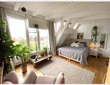 Helles Schlafzimmer in einem Dachgeschoss mit Holzbalkendecke, einem großen Bett mit grauer Tagesdecke und Kissen, vielen Zimmerpflanzen und einem niedrigen Holztisch mit weiteren Pflanzen. Große Fenster bieten Blick auf Häuser und Bäume draußen.