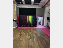 Open studio or event space with a wood plank floor and a pink carpet runway leading to a backdrop with rainbow light streak graphics in front of black curtains. The ceiling has exposed pipes and acoustic panels, and the stage area includes balloons, a large “7” balloon, a tinsel photo curtain, and a speaker on a stand.