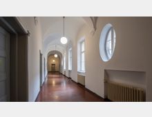 Ein langer, schmaler Flur mit weiß gestrichenen Wänden, hohen Decken mit Gewölbebögen und hängenden kugelförmigen Leuchten. Auf der rechten Seite befinden sich Fenster, darunter kreisrunde und rechteckige, sowie Heizkörper; der Boden ist aus roten Fliesen.