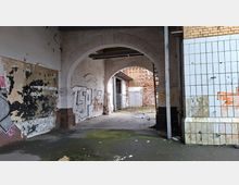 Verlassene Industriehalle mit großen gemauerten Rundbögen, abblätterndem Putz und Graffiti an den Wänden. Rechts sind weiß gekachelte Wandflächen zu sehen; im Hintergrund führen die Bögen in einen offenen Bereich mit freiliegendem Mauerwerk.