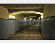 Ein geschwungener, leicht beleuchteter Fußgängertunnel mit blau gefliesten Wänden und einer Decke mit gewölbten Betonträgern. An den Wänden befinden sich regelmäßig angeordnete rechteckige Lampen.