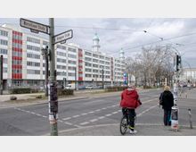 Straßenecke an der Kreuzung Warschauer Straße und Boxhagener Straße in Berlin, mit einem modernen, weiß-roten Mehrstöckigen Gebäude im Hintergrund und leicht erkennbaren grünen Kuppeln. Im Vordergrund sind Verkehrszeichen, Fußgänger, ein Fahrradfahrer und Plakate an einem Mast zu sehen, während Autos am Straßenrand parken.
