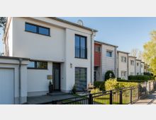 Moderne Reihenhäuser mit minimalistischer Architektur, weißer Fassade und großen Fenstern, umgeben von kleinen gepflegten Vorgärten. Vor den Häusern verläuft ein Gehweg mit einem Metallzaun, dahinter stehen Hecken und Sträucher.