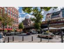 Straßenszene in einer Stadt mit mehrstöckigen Backstein- und Putzfassaden, darunter Geschäfte und Cafés. Im Hintergrund verläuft eine erhöhte Bahntrasse, auf der eine rote S-Bahn fährt, und die Straße ist von Autos und Fußgängern belebt.