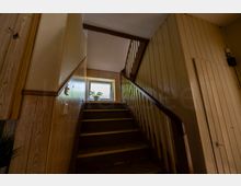 Innenaufnahme eines schmalen Treppenhauses mit Holzvertäfelung und dunkel gestrichener Holztreppe mit Geländer, die zu einem Podest führt. Am Ende des Treppenlaufs befindet sich ein Fenster mit einer Topfpflanze auf der Fensterbank, seitlich sind Türen und eine Wandleuchte zu sehen.