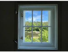 Blick durch ein modernes, weiß gerahmtes Sprossenfenster auf eine grüne Landschaft mit Bäumen und einem Fluss unter blauem Himmel.