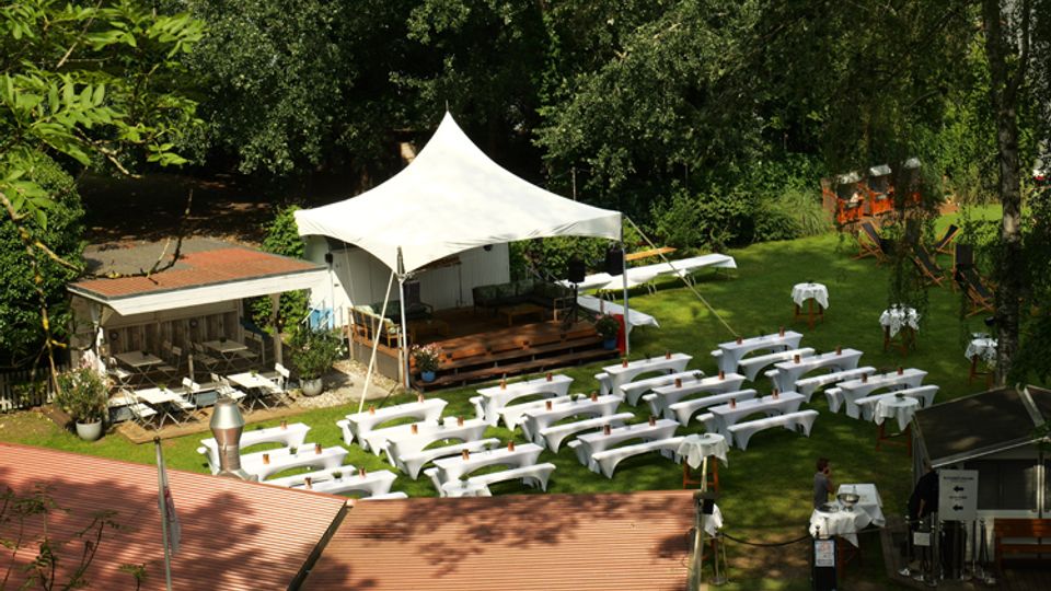 Gartenanlage mit Rasenfläche, auf der zahlreiche lange Tische und Bänke mit weißen Hussen in Reihen stehen. Im Hintergrund befindet sich eine kleine Bühne unter einem weißen Zeltdach, umgeben von Bäumen und kleinen Nebengebäuden.